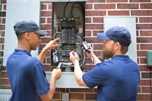 Two men electricians working on a residential breaker box.  They use tools to assess the repair and wear matching blue uniforms.  The multi-ethnic group is discussing next steps in the job repair.  Electric meter to side.