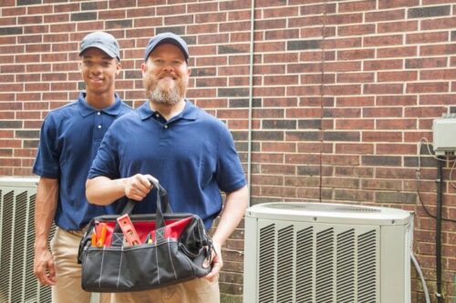 Multi-ethnic team of men repairing a home's air conditioner unit outdoors. They have completed the repairs.  They both wear blue uniforms and man in foreground holds a tool bag.